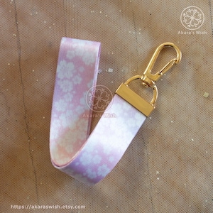 May include: A floral patterned wrist strap in shades of pink and white, attached to a gold-colored metal clasp. The strap features a repeating flower design. The Etsy shop name "Akaras Wish" is visible on the strap.