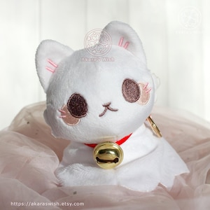 May include: A white plush cat with brown eyes, pink accents, and a red collar with a gold bell. The cat has a smiling face and is sitting on a pink surface. The Etsy shop URL is visible at the bottom.