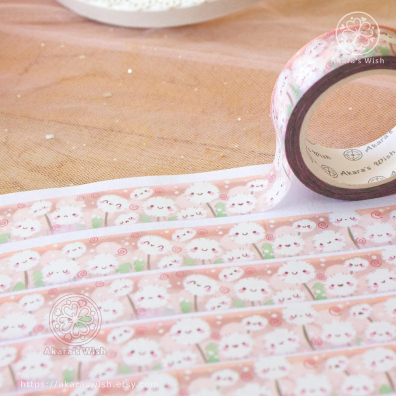 May include: A roll of pink and white washi tape with a repeating pattern of cartoon dandelions with smiling faces. The tape is labelled "Akara's Wish" and has a website address printed on the bottom.