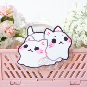 May include: A clear sticker depicting two cartoon white cats with black eyes and pink cheeks. The sticker is placed on a pink plastic crate, with flowers in the background. The text "Akara's Wish" is visible on the sticker.