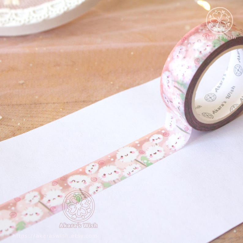 May include: Pink and white washi tape with a repeating pattern of cartoon dandelions with faces. The tape is unrolled on a white surface and the roll of tape is visible in the background. The tape has the text "Akara's Wish" on it.