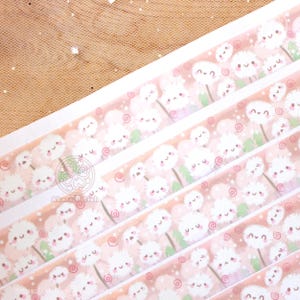 May include: A roll of washi tape featuring a repeating pattern of white dandelions with pink cheeks and green stems on a light pink background. The dandelions have small black eyes and a smiling mouth. The tape is decorated with small pink swirls.