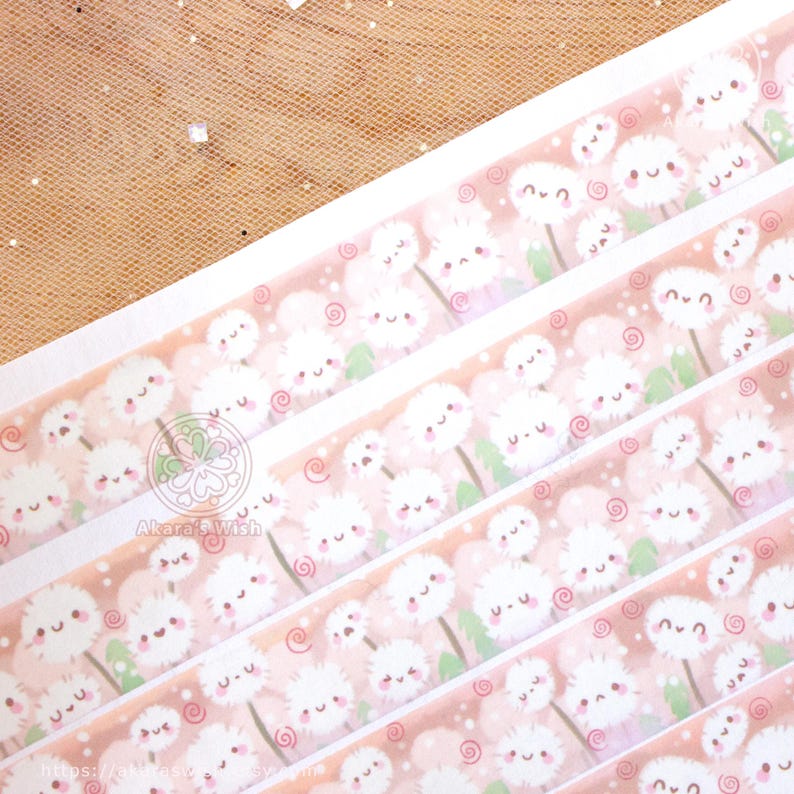 May include: A roll of washi tape featuring a repeating pattern of white dandelions with pink cheeks and green stems on a light pink background. The dandelions have small black eyes and a smiling mouth. The tape is decorated with small pink swirls.