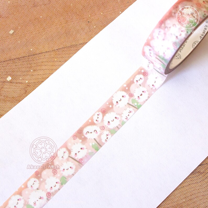 May include: A roll of washi tape with a pink and white design featuring cute cartoon dandelions with faces. The tape is unrolled on a white surface and the roll is visible in the top right corner of the image. The text "Akara's Wish" is visible on the roll of tape.