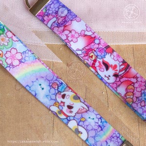 May include: Two colorful keychains with a white background. One keychain features a white cat with pink and purple flowers. The other keychain features a white fox with pink and purple flowers. Both keychains have a rainbow design.