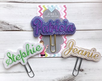 May include: Three personalized name paper clips. The names "Patricia" in purple, "Stehpie" in green, and "Jeanie" in gold are embroidered on white felt. Each clip is attached to a gold-colored paper clip, with a decorative backing.