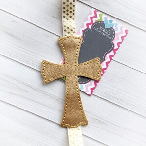 May include: A gold leather cross bookmark with a white ribbon with gold polka dots. The bookmark is attached to a business card that says "Noelle's Custom Creations".