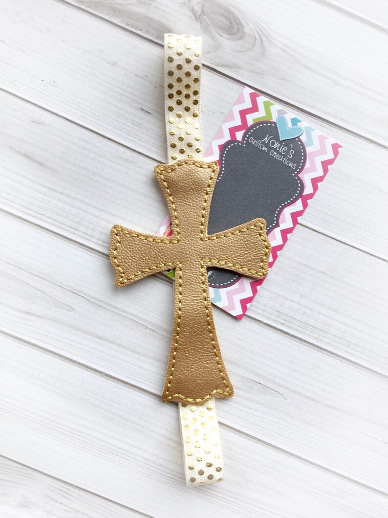 May include: A gold leather cross bookmark with a white ribbon with gold polka dots. The bookmark is attached to a business card that says "Noelle's Custom Creations".