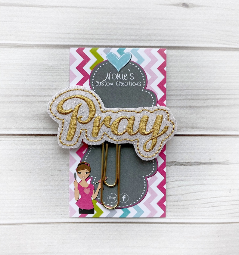 May include: A gold glitter paper clip with the word "Pray" embroidered in gold thread. The paper clip is attached to a gray card with a pink, white, and blue chevron pattern. The card also features a small heart and the text "Nonie's custom creations".