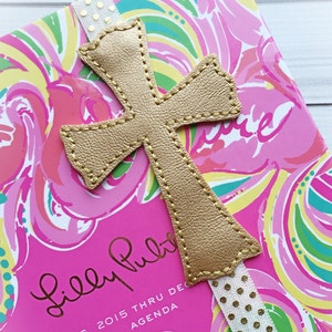 May include: Gold leather cross bookmark with a white ribbon with gold polka dots. The bookmark is on a pink and green patterned Lilly Pulitzer agenda.