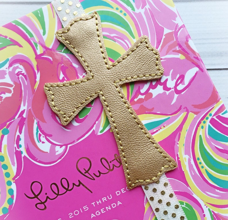 May include: Gold leather cross bookmark with a white ribbon with gold polka dots. The bookmark is on a pink and green patterned Lilly Pulitzer agenda.