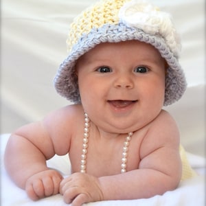 May include: A baby wearing a yellow and grey crocheted hat with a white flower on top and a pearl necklace. The baby is smiling and looking at the camera.