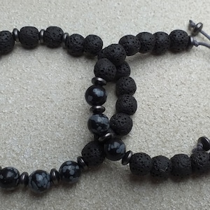 May include: Two beaded bracelets, one black lava rock and one with black lava rock and grey and white speckled beads. Both bracelets have a grey cord with a knot at the end.