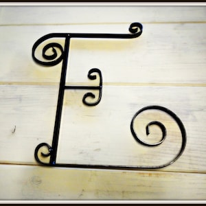 May include: Black metal letter F with decorative swirls. The letter is mounted on a white wooden surface.
