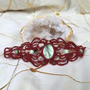 Puede incluir: Una pulsera de macramé de color rojo oscuro con una piedra preciosa de labradorita ovalada en el centro. El intrincado diseño de cordón anudado está acentuado con pequeñas cuentas redondas en tonos verdes y beige, creando un patrón detallado.