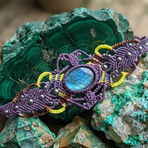 Puede incluir: Una pulsera de macramé con una piedra central ovalada de labradorita con iridiscencia azul. La pulsera está hecha de cordón morado con detalles en hilo amarillo y color cobre, decorada con pequeñas cuentas metálicas plateadas.