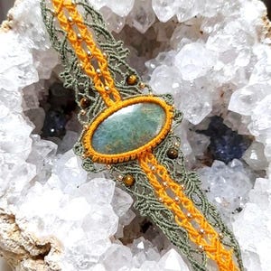 Könnte beinhalten: Ein Makramee-Armband mit einem zentralen ovalen grünen Stein, eingefasst in leuchtend gelbe Kordel, flankiert von olivgrünen Knotenmustern, akzentuiert mit kleinen Metallperlen und runden braunen Steinen im gesamten komplexen Design.