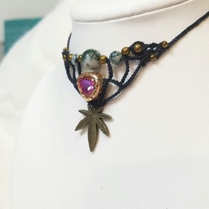 May include: A handmade macrame choker necklace. The black cord supports green and gold beads, a heart-shaped pink gem, and a bronze-colored cannabis leaf pendant. The necklace is displayed on a white mannequin.
