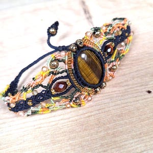 May include: A macrame bracelet featuring a central oval tiger's eye gemstone surrounded by dark blue, orange, and yellow woven threads. The design includes small metallic gold beads and faceted brown beads integrated into the intricate knotwork pattern.