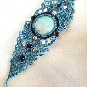 Puede incluir: Una pulsera de macramé color verde azulado con una piedra grande, redonda y de color verde pálido en el centro. El intrincado diseño de cordón anudado está acentuado con pequeñas cuentas negras, espaciadores metálicos cuadrados plateados y una cuenta plateada texturizada.