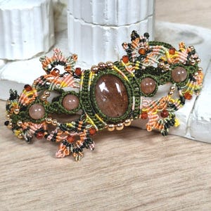 Peut inclure: Barrette à cheveux macramé faite à la main. Elle présente une grande pierre précieuse ovale marron au centre, entourée de petites pierres précieuses roses rondes. La barrette a un design détaillé avec des nœuds macramé verts, oranges et noirs et des perles dorées.