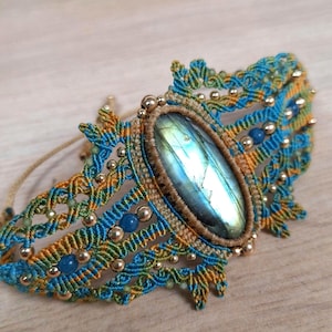 May include: A handcrafted macrame bracelet featuring a large, oval labradorite stone. The bracelet is intricately woven with blue, gold, and green threads, accented with small gold beads and blue gemstone beads. The design is symmetrical and detailed.