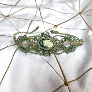 May include: A macrame bracelet featuring a central oval labradorite gemstone with iridescent yellow and blue flashes. The sage green cord is intricately knotted with small metallic gold beads and tiny green glass seed beads throughout the design.