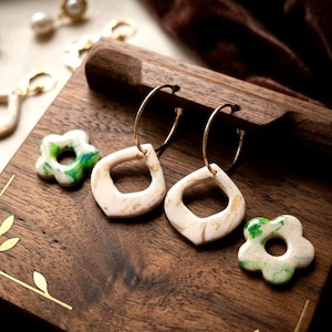 May include: A wooden display holds two pairs of earrings. One pair features gold hoops with cream-colored, geometric pendants with gold flecks. Two flower-shaped earrings with green and white swirls are also present.