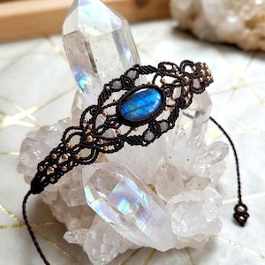 Puede incluir: Una pulsera de macramé marrón oscuro con una piedra preciosa de labradorita de forma ovalada con iridiscencia azul en el centro. El diseño de cordón trenzado está acentuado con pequeñas cuentas metálicas y cordones ajustables con cuentas.