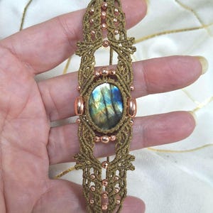 May include: A handmade macrame bracelet featuring a central oval labradorite stone with blue, green, and yellow iridescence. The bracelet is crafted with tan-colored cord and accented with copper-colored beads and oval embellishments.
