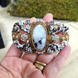 Puede incluir: Una pulsera de macramé ancha con un gran cabujón de piedra lunar ovalado e iridiscente en el centro. La banda está hecha de cordón marrón, negro y blanco, decorada con cuentas de metal plateado en forma de flor, pequeños cristales y cuentas redondas.