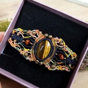 Op de afbeelding: Handgemaakte macramé armband met een grote ovale tijgeroogsteen in het midden, omringd door ingewikkelde knoopwerk in marineblauwe, oranje, gele en roze draden. Geaccentueerd met kleine gouden kralen en gepresenteerd in een roze sieradendoos.