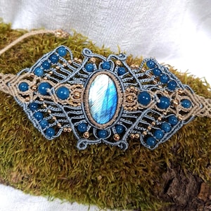 May include: A macrame bracelet featuring a central oval labradorite gemstone with blue iridescence, surrounded by intricate grey cord knotting, small round blue beads, and tiny metallic gold-toned beads arranged in a symmetrical pattern.