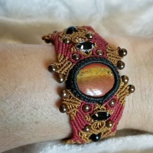 May include: A handmade macramé bracelet featuring a large oval stone with red, yellow, and brown hues. The bracelet has a red and tan woven design, accented with black stones and bronze-coloured beads.
