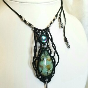 Peut inclure: Un collier macramé fait à la main avec une grosse pierre en forme de goutte d'eau aux teintes vertes et dorées. Le pendentif est orné d'une pierre irisée et de perles argentées. Le collier a un cordon noir et une pointe argentée en bas.
