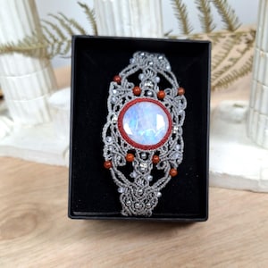 Peut inclure: Un pendentif de collier en macramé fait à la main avec une grande pierre précieuse ovale irisée au centre. Le pendentif est gris avec des accents rouges et orange, présenté dans une boîte noire. La pierre précieuse est bordée de rouge.