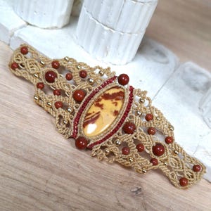 Peut inclure: Un bracelet en macramé avec une pierre centrale ovale aux motifs bruns et jaunes, entourée de macramé rouge et beige. De petites perles rouges et dorées sont incorporées. Le bracelet est présenté sur une surface en bois.