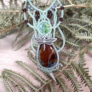 Op de afbeelding: Handgemaakte macramé ketting met een grote, druppelvormige bruin en blauw agaat stenen hanger. De ketting heeft een lichtgroen koord met decoratieve kralen en een zilveren libellenbedel. De hanger is ongeveer 5 cm lang.