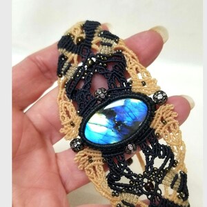 Peut inclure: Bracelet en macramé orné d'une pierre de labradorite ovale, aux reflets bleus et verts. Le bracelet est réalisé avec du cordon macramé noir et beige, agrémenté de petites perles et d'éléments décoratifs.
