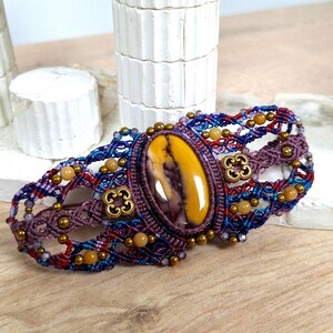Peut inclure: Bracelet en macramé fait à la main, orné d'une pierre ovale jaune et violette. Le bracelet combine des fils bleus, rouges et violets, avec des perles dorées et des éléments métalliques décoratifs. Il est présenté sur une surface en bois.