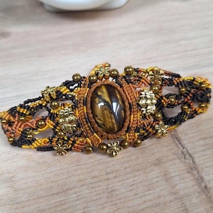 Könnte beinhalten: Ein Makramee-Armband mit einem ovalen Tigeraugenstein in der Mitte. Das Armband ist mit schwarzen, gelben und orangefarbenen Fäden gewebt und mit kleinen goldfarbenen Blumen- und Perlenverzierungen versehen. Es wird auf einer hellen Holzoberfläche präsentiert.