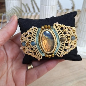 Könnte beinhalten: Ein Makramee-Armband mit einem zentralen, ovalen Labradorit-Edelstein, der irisierende blaue und goldene Reflexe aufweist. Das Stück ist aus beigefarbenem Garn, türkisfarbenen Akzenten, kleinen goldenen Metallperlen sowie grauen Kristallperlen gefertigt.