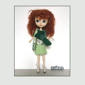 Può includere: Bambola con capelli ricci rossi, che indossa un completo verde, tra cui una gonna a pieghe, un maglione e una borsa con un motivo floreale bianco. La bambola indossa scarpe da ginnastica beige. La parola "art'co" è visibile.