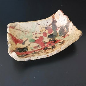 Puede incluir: Un plato rectangular de cerámica con un borde irregular ligeramente ondulado. El plato está decorado con un esmalte moteado en tonos de marrón, rojo y verde.