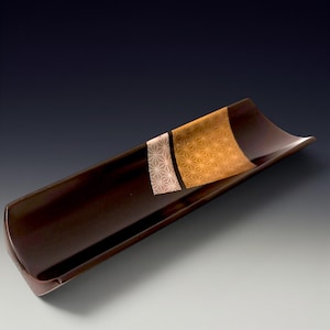 Puede incluir: Una fuente rectangular larga de color marrón oscuro con lados suavemente curvados. Una sección decorativa presenta un patrón geométrico dorado y blanco. La fuente es probablemente de cerámica o madera lacada, adecuada para servir sushi o aperitivos.