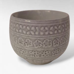 May include: A small, gray ceramic bowl with a rounded shape. The bowl features a textured floral pattern around the exterior. The rim is smooth, and the bowl has a slightly uneven surface, giving it a rustic appearance. The bowl is suitable for serving small portions.