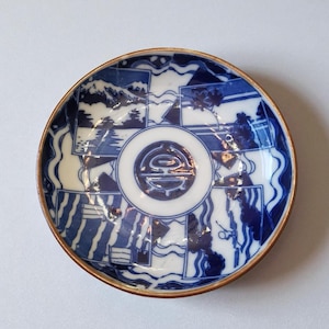 May include: A round, blue and white ceramic plate with a brown rim. The plate features a central design and surrounding landscape scenes. The design includes mountains, trees, and water. The plate is approximately 6 inches in diameter.