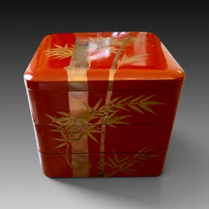 May include: A square, three-tiered stackable box with a glossy red finish. The exterior features a decorative gold-toned bamboo stalk design that spans across all three levels, including the lid, with detailed leaves extending outward.