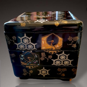 Puede incluir: Una caja bento japonesa lacada en negro con un diseño en capas. La caja presenta detalles dorados y de nácar, incluyendo motivos de grullas y florales. La caja tiene un acabado brillante y una forma rectangular.