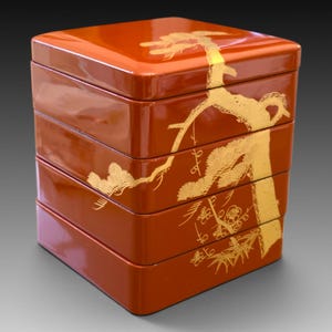Op de afbeelding: Een vierkante, vierlaagse stapelbare doos met een glanzende, diep oranje-rode afwerking. De buitenkant is voorzien van een doorlopend, gedetailleerd goudkleurig grafisch ontwerp dat een gestileerde dennentak over alle vier de niveaus weergeeft.